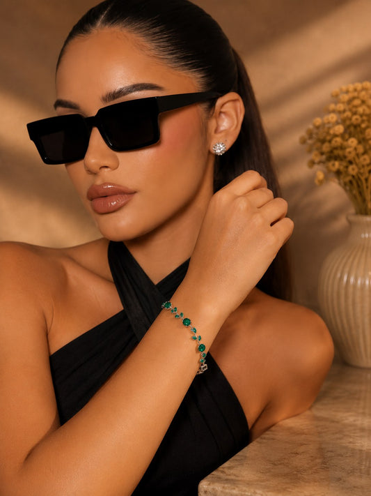 A LUSHÉ vine bracelet with emerald-green stones, worn with black square sunglasses